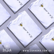 Футболка бджілка це життя. Біла футболка з коротким рукавом, зображена з правого ракурсу на манекені. На футболці є принт у вигляді бджілки з жовто-чорними смужками та написом 'Ж.ж.ж.ж.' під нею. Фон світлий, з легким градієнтом.