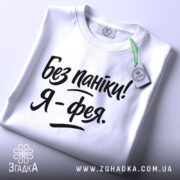 Футболка без паніки я фея, білого кольору, складена на поверхні, фон світло-сірий, принт чорного кольору з написом 'Без паніки! Я - фея.', на комірці видно етикетку з зеленим шнурком.