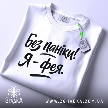 Футболка без паніки я фея, білого кольору, складена на поверхні, фон світло-сірий, принт чорного кольору з написом 'Без паніки! Я - фея.', на комірці видно етикетку з зеленим шнурком.