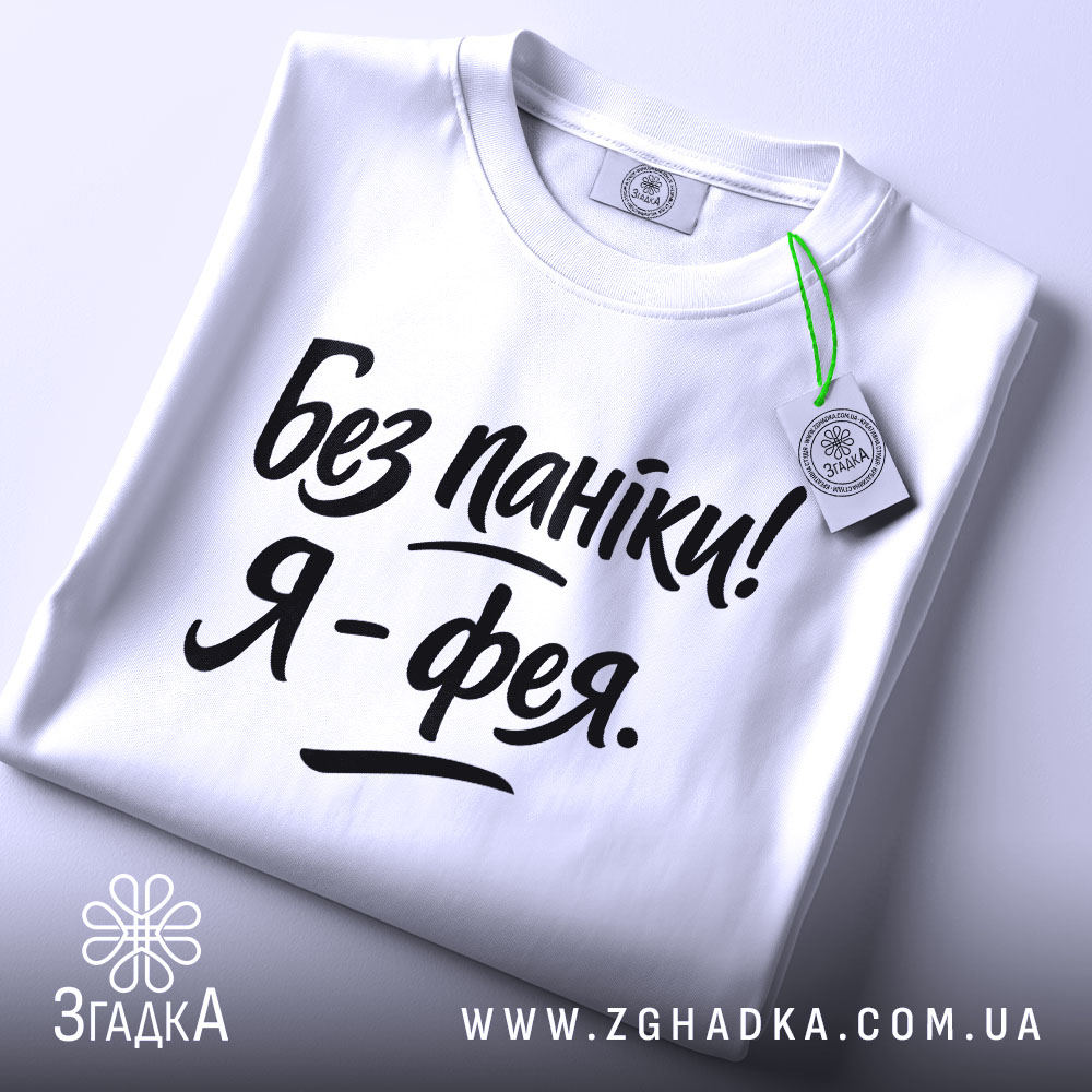 Футболка без паніки я фея, білого кольору, складена на поверхні, фон світло-сірий, принт чорного кольору з написом 'Без паніки! Я - фея.', на комірці видно етикетку з зеленим шнурком.