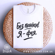 Футболка без паніки я фея, білого кольору, складена на плетеній кошику, фон світло-сірий, принт чорного кольору з написом 'Без паніки! Я - фея.'