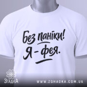 Футболка без паніки я фея, білого кольору, складена на поверхні, фон світло-сірий, принт чорного кольору з написом 'Без паніки! Я - фея.', на комірці видно етикетку з зеленим шнурком.