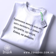 Футболка біль художника, біла, зображена зверху, на столі, фон світло-сірий, принт чорного кольору з текстом про художників, з зеленим ярликом.