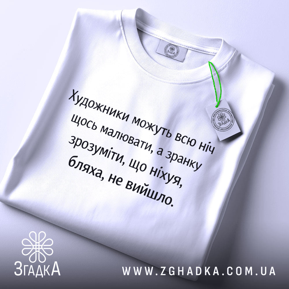 Футболка біль художника, біла, зображена зверху, на столі, фон світло-сірий, принт чорного кольору з текстом про художників, з зеленим ярликом.