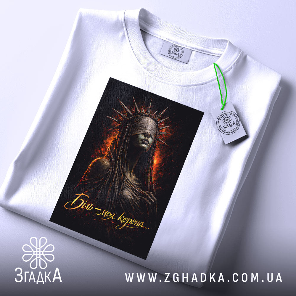 Футболка біль моя корона. Біла футболка, складена на поверхні, з принтом темної жінки в короні, фон з вогняними відтінками, на комірці видна етикетка.