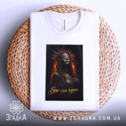 Футболка біль моя корона. Біла футболка, складена на плетеній основі, з принтом темної жінки в короні, фон з вогняними елементами, етикетка видна на комірці.