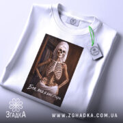 Футболка бля я стара, біла, складена на поверхні, фон світлий, принт з скелетом у рушнику, текст під принтом, на футболці є етикетка з зеленим шнурком.