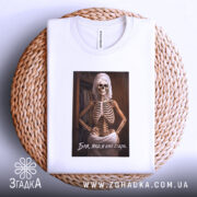 Футболка бля я стара, біла, складена на плетеній кошику, фон світлий, принт з скелетом у рушнику, текст під принтом.