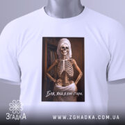 Футболка бля я стара, біла, складена на поверхні, фон світлий, принт з скелетом у рушнику, текст під принтом, на футболці є етикетка з зеленим шнурком.