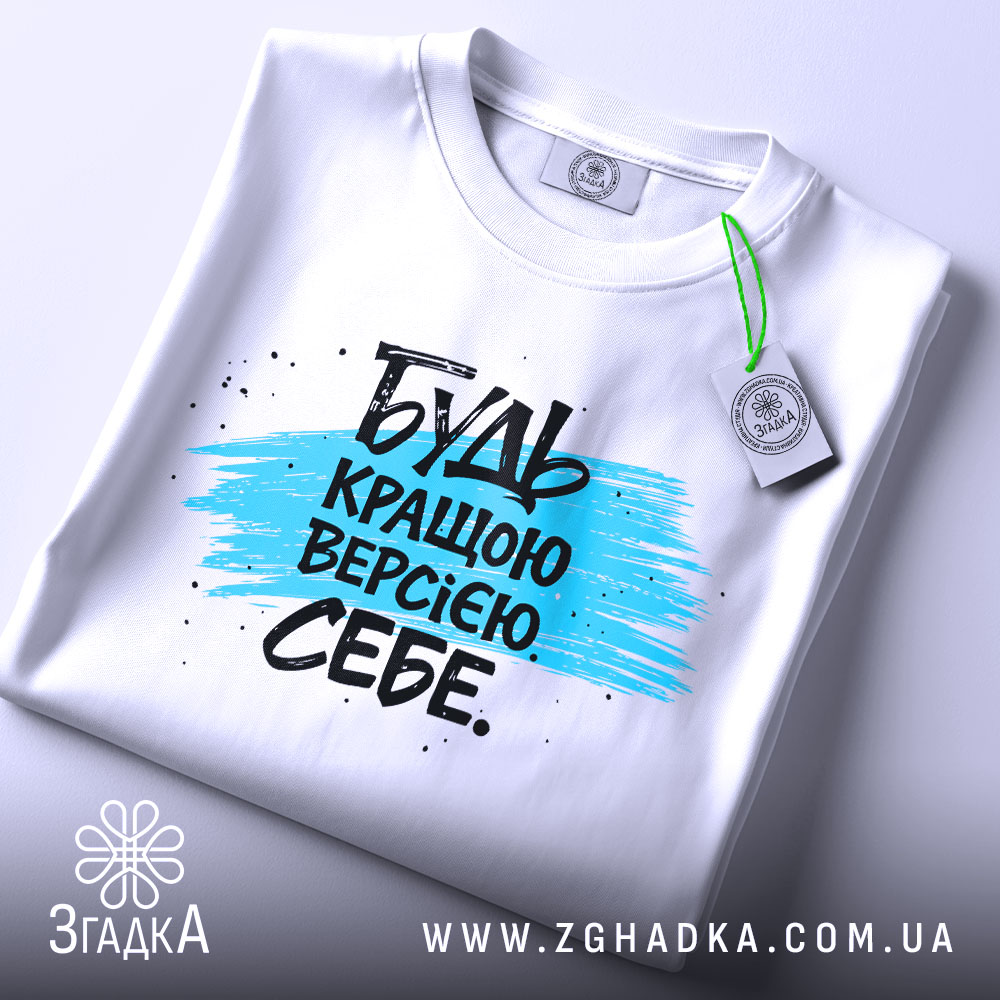 Футболка будь кращою версією на столі, білого кольору, з принтом у вигляді синьої смуги та чорного тексту, з етикеткою зеленого кольору, фон світло-сірий.