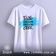 Футболка будь кращою версією на вішалці, білого кольору, з принтом у вигляді синьої смуги та чорного тексту, фон світло-сірий.