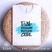 Футболка будь кращою версією на плетеній основі, білого кольору, з принтом у вигляді синьої смуги та чорного тексту, фон світло-сірий.