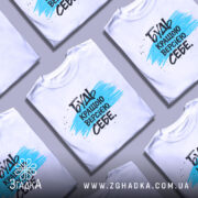 Футболка будь кращою версією на манекені, білого кольору, з принтом у вигляді синьої смуги та чорного тексту, фон світло-сірий.