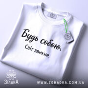 Футболка будь собою, білого кольору, складена на поверхні, фон світло-сірий, принт чорного кольору з написом 'Будь собою. Світ звикне.', на комірці є етикетка.