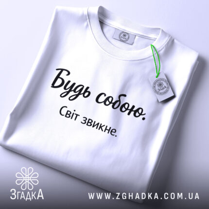 Футболка будь собою, білого кольору, складена на поверхні, фон світло-сірий, принт чорного кольору з написом 'Будь собою. Світ звикне.', на комірці є етикетка.