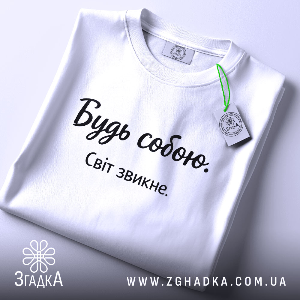 Футболка будь собою, білого кольору, складена на поверхні, фон світло-сірий, принт чорного кольору з написом 'Будь собою. Світ звикне.', на комірці є етикетка.