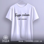 Футболка будь собою, білого кольору, підвішена на вішалці, фон світло-сірий, принт чорного кольору з написом 'Будь собою. Світ звикне.'