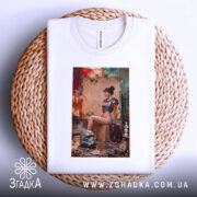 Футболка бунтарка стімпанк, біла, складена на плетеній основі, зображення моделі сидить на коробці в стімпанк-інтер'єрі, фон з червоними і зеленими елементами, деталі принта включають шолом і годинник на підлозі.