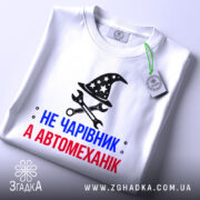 Футболка чарівник автомеханік, біла, зображена зверху, фон світлий, принт з магічним капелюхом та інструментами, текст червоний і синій, на футболці є етикетка.