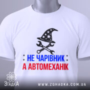 Футболка чарівник автомеханік, біла, зображена зверху, фон світлий, принт з магічним капелюхом та інструментами, текст червоний і синій, на футболці є етикетка.