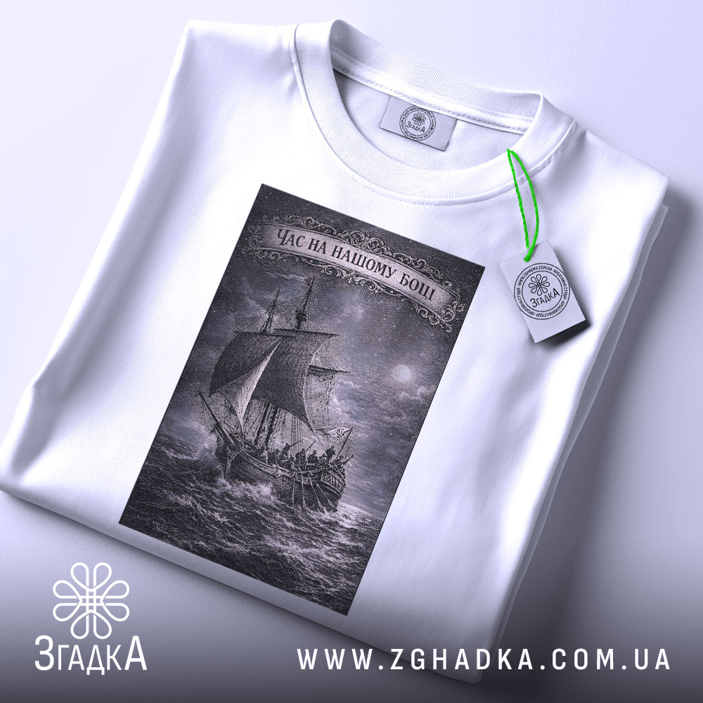 Футболка час на нашому боці. Складена біла футболка з принтом, зображення корабля на фоні бурного моря, ракурс зверху, фон світлий, деталі принта виконані в сірому кольорі, на футболці є етикетка з зеленим шнурком.