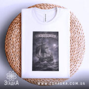 Футболка час на нашому боці. Складена біла футболка з принтом, зображення корабля на фоні бурного моря, ракурс зверху, фон світлий, деталі принта виконані в сірому кольорі, футболка лежить на плетеній основі.