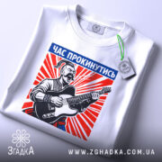 Футболка час прокинутись. Біла футболка, складена на столі, з принтом чоловіка з гітарою. Принт яскравий, з червоними променями, текст "ЧАС ПРОКИНУТИСЬ" розташований у верхній частині. На футболці є етикетка з зеленим шнурком.