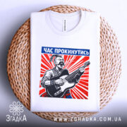 Футболка час прокинутись. Біла футболка, складена на плетеній основі, з принтом чоловіка з гітарою. Принт яскравий, з червоними променями, текст "ЧАС ПРОКИНУТИСЬ" розташований у верхній частині.