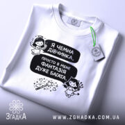 Футболка чемна дівчинка, білого кольору, зображена зверху, на фоні світлого кольору, принт з текстом і малюнками ангела та дівчинки з чортом, з зеленим ярликом.