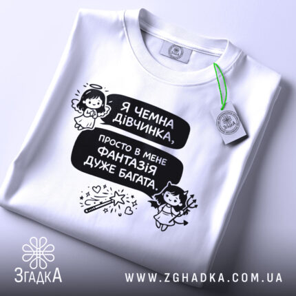 Футболка чемна дівчинка, білого кольору, зображена зверху, на фоні світлого кольору, принт з текстом і малюнками ангела та дівчинки з чортом, з зеленим ярликом.