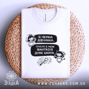 Футболка чемна дівчинка, білого кольору, зображена на плетеній основі, фон світлий, принт містить текст і малюнки ангела та дівчинки з чортом.