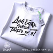 Футболка дай Боже кожному, біла, складена на поверхні, фон світло-сірий, принт чорного кольору з текстом 'Дай Боже кожний Такого, як я!' у стилі рукописного шрифту, з етикеткою зеленого кольору.