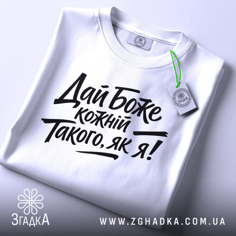 Футболка дай Боже кожному, біла, складена на поверхні, фон світло-сірий, принт чорного кольору з текстом 'Дай Боже кожний Такого, як я!' у стилі рукописного шрифту, з етикеткою зеленого кольору.