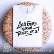Футболка дай Боже кожному, біла, складена на плетеній кошику, фон світло-сірий, принт чорного кольору з текстом 'Дай Боже кожний Такого, як я!' у стилі рукописного шрифту.