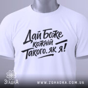 Футболка дай Боже кожному, біла, складена на поверхні, фон світло-сірий, принт чорного кольору з текстом 'Дай Боже кожний Такого, як я!' у стилі рукописного шрифту, з етикеткою зеленого кольору.