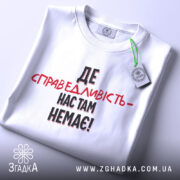Футболка де справедливість, біла, складена на столі, фон світло-сірий, принт чорного та червоного кольорів з текстом 'ДЕ СПРАВЕДЛИВІСТЬ - НАС ТАМ НЕМЕЄ!', з етикеткою на зеленій стрічці.