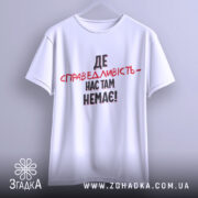 Футболка де справедливість, біла, підвішена на вішалці, фон світло-сірий, принт чорного та червоного кольорів з текстом 'ДЕ СПРАВЕДЛИВІСТЬ - НАС ТАМ НЕМЕЄ!'.