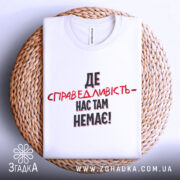 Футболка де справедливість, біла, складена на плетеній кошику, фон світло-сірий, принт чорного та червоного кольорів з текстом 'ДЕ СПРАВЕДЛИВІСТЬ - НАС ТАМ НЕМЕЄ!'.