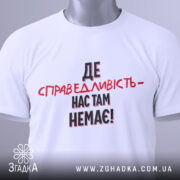 Футболка де справедливість, біла, складена на столі, фон світло-сірий, принт чорного та червоного кольорів з текстом 'ДЕ СПРАВЕДЛИВІСТЬ - НАС ТАМ НЕМЕЄ!', з етикеткою на зеленій стрічці.