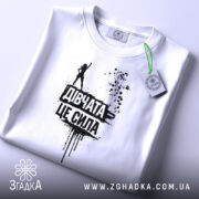 Футболка дівчата це сила, білого кольору, зображена зверху, складена на поверхні, фон світлий, принт чорного кольору з написом 'Дівчата це сила' та силуетом дівчини, на футболці є етикетка з зеленим шнурком.