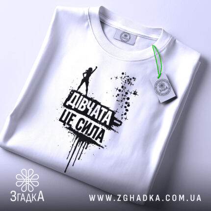 Футболка дівчата це сила, білого кольору, зображена зверху, складена на поверхні, фон світлий, принт чорного кольору з написом 'Дівчата це сила' та силуетом дівчини, на футболці є етикетка з зеленим шнурком.