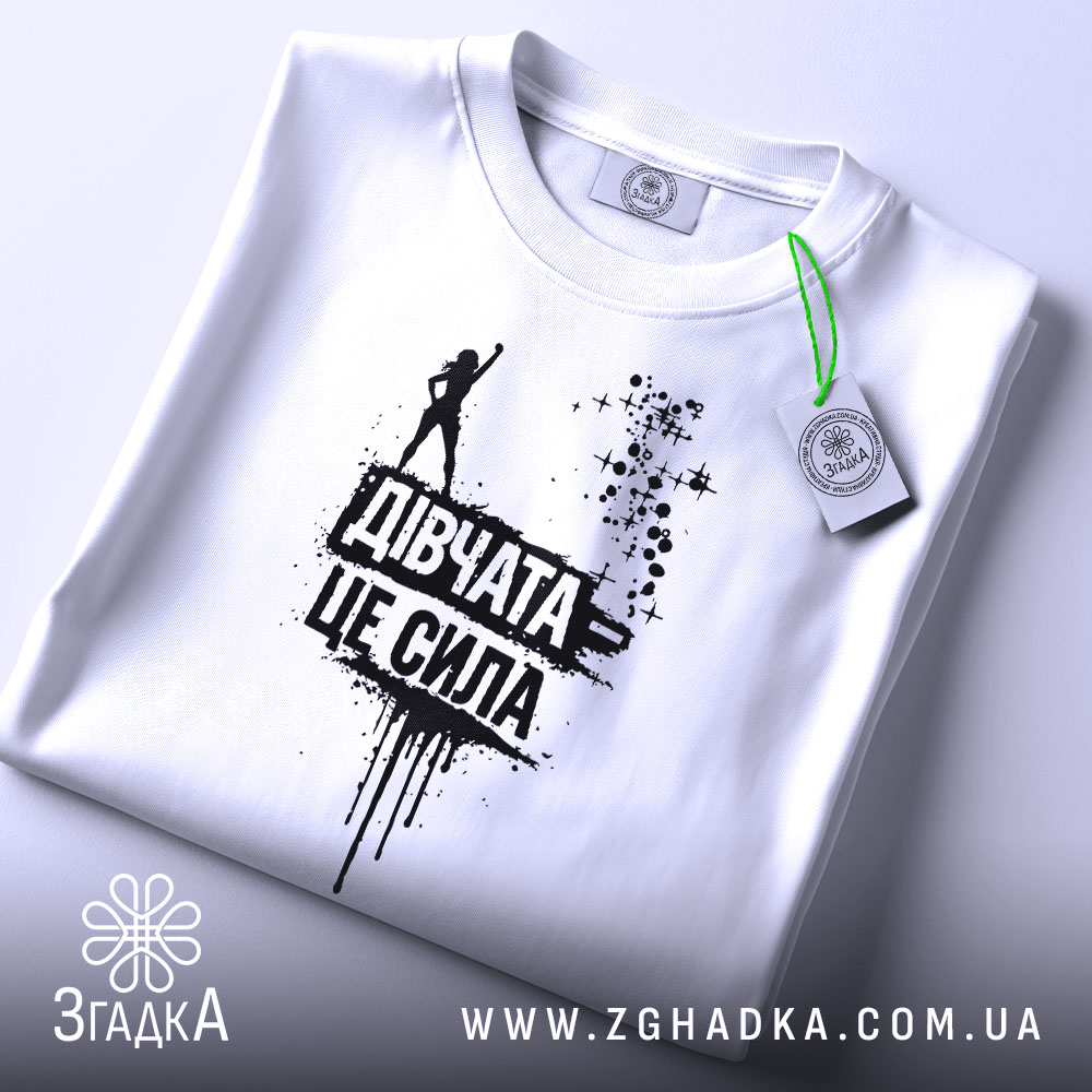 Футболка дівчата це сила, білого кольору, зображена зверху, складена на поверхні, фон світлий, принт чорного кольору з написом 'Дівчата це сила' та силуетом дівчини, на футболці є етикетка з зеленим шнурком.