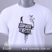 Футболка дівчата це сила, білого кольору, зображена зверху, складена на поверхні, фон світлий, принт чорного кольору з написом 'Дівчата це сила' та силуетом дівчини, на футболці є етикетка з зеленим шнурком.