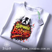 Футболка дівчина вогонь, біла, зображена зверху на столі, фон світлий, принт з написом 'Дівчина Вогонь' в червоних і жовтих кольорах, оточений вогняними ефектами, з зеленим ярликом.