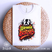 Футболка дівчина вогонь, біла, зображена зверху на плетеному кошику, фон світлий, принт з написом 'Дівчина Вогонь' в червоних і жовтих кольорах, оточений вогняними ефектами.