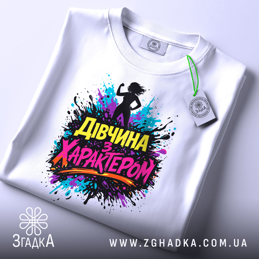 Футболка дівчина з характером, білого кольору, розкладена на поверхні, фон світлий, принт з силуетом дівчини та кольоровими сплесками фарби, текст "Дівчина з характером" яскравих кольорів, на комірці видно етикетку.