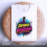 Футболка дівчина з характером, білого кольору, складена на плетеній кошику, фон світлий, принт з силуетом дівчини та кольоровими сплесками фарби, текст "Дівчина з характером" яскравих кольорів.