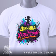 Футболка дівчина з характером, білого кольору, розкладена на поверхні, фон світлий, принт з силуетом дівчини та кольоровими сплесками фарби, текст "Дівчина з характером" яскравих кольорів, на комірці видно етикетку.