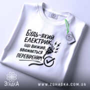 Футболка для електрика, білого кольору, складена на поверхні, фон світло-сірий, принт чорного кольору з текстом і малюнком електричної вилки, на футболці є етикетка.