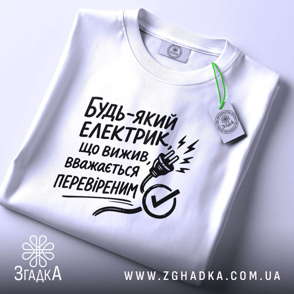 Футболка для електрика, білого кольору, складена на поверхні, фон світло-сірий, принт чорного кольору з текстом і малюнком електричної вилки, на футболці є етикетка.